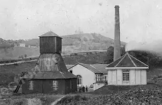 Photo en noir et blanc de petits bâtiments, avec grande cheminée et tour pyramidale en bois fermée au pied de la colline de la chapelle Notre-Dame-du-Haut.