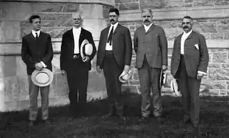 Photographie en noir et blanc de cinq hommes en costume posant chapeau à la main