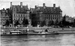 Photographie en noir et blanc montrant une vue d'ensemble du bâtiment, pris de l'autre côté de la rivière qu'on voit au premier plan.