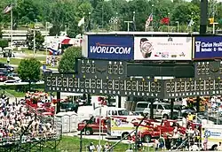 Phtographie en couleur d'un paddock de course, avec des spectateurs à gauche, et un tableau des scores au centre, surmonté de plusieurs drapeaux.