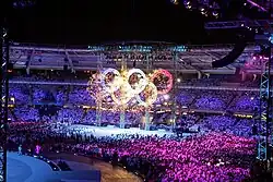 Photographie d'un spectacle de nuit dans un stade. La foule est illuminée en rose et violet, les anneaux olympiques illuminés par pyrotechnie au centre du terrain.