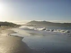 Photo en couleur d'une longue plage avec des vagues d'écume et des montagnes au loin, dans la lumière du matin.
