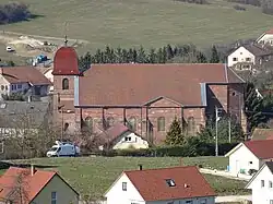 Église en grès rose de profil. Successivement : un clocher comtois en tuiles vernissées rouges, chevet sans transept et nef.