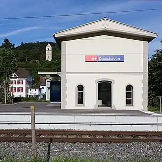 Photo avec en premier plan les rails, le quai et la façade du bâtiment. À l'arrière-plan à gauche des bâtiments du village et un clocher au milieu des arbres sur une colline. le ciel est bleu.