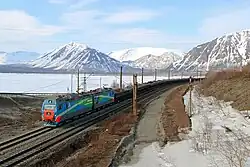 Un convoi de wagons noirs remplis de charbon occupe le centre de l'image, tiré par une locomotive diesel. La voie ferrée, à double voie, longe les rives d'un lac, au centre de l'image, aux eaux gris-bleu qui reflètent un ciel chargé de nuages. À droite, la berge rocheuse lac est dénudée de végétation haute, seulement des herbes. En arrière-plan, des montagnes haute et pelées, partiellement enneigées, ferment l'horizon.
