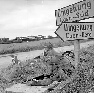 Photo en noir et blanc de deux soldats à couvert, en position dans un fossé près d'une route, sous un panneau routier écrit en allemand et indiquant la direction de Caen.