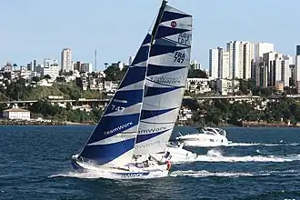 Voilier bleu et blanc dont on remarque l'avant arrondi, lancé sur l'océan, suivi par deux vedettes. À l'arrière-plan, une ville et ses gratte-ciel.