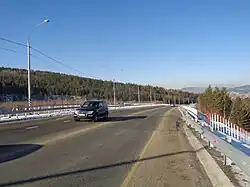 Photographie depuis le bas-côté d'une route avec une voie de chaque côté et une voiture au centre. De la taïga se trouve derrière la rue.