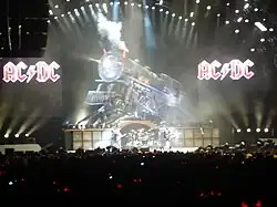 Quatre membre de groupe sur scène devant un public, avec une locomotive factice et deux logos AC/DC en fond.