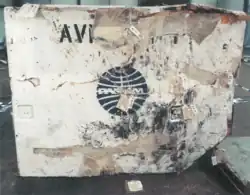 Photo d’un conteneur à bagages blanc fortement endommagé, portant le logo bleu de Pan Am. Les parois sont déformées, marquées par des traces d’impact et de brûlure. Les lettres AV restent partiellement visibles sur la partie supérieure gauche.