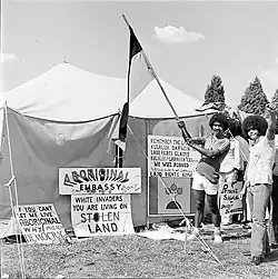 Photo noir et blanc de 3 aborigènes devant une tente décorée de slogans