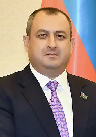 Photographie d'un homme en costume, l'air sérieux, portant une broche aux couleurs de l'Azerbaïdjan, devant un drapeau azerbaïdjanais.