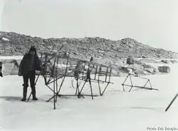 Photo en noir et blanc d'un squelette métallique d'avion posé sur la neige, une personne postant devant.