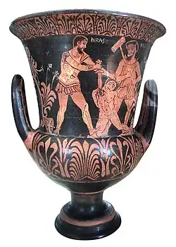 Calice de couleur noire et ocre avec deux anses sur lequel est représenté soldat plantant un glaive dans un homme nu agenouillé au sol, regardés par un autre homme avec un marteau.