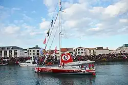 Voilier rouge de profil. Sur le quai, une foule considérable.