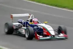 Photographie d'une monoplace de Formule 3 blanche, bleu foncé, rouge, verte, vue de trois-quarts droit avec un flou artistique sur la photo. Seule la zone autour du casque est nette.