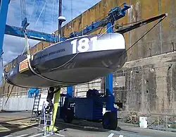 Porté par un élévateur, bateau bleu numéro 181 vu en contre-plongée. Perché sur un escabeau, un homme travaille sur la fixation de la quille.