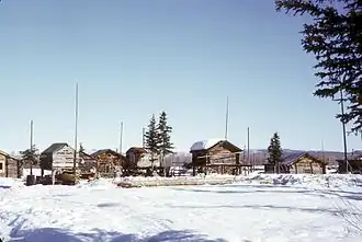 La photo représente un village de petites maisons de bois, certaines sur pilotis, au milieu de la neige.
