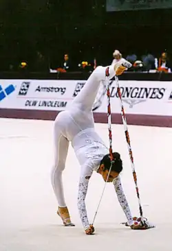Photo d'une gymnaste faisant une figure avec un ruban, vêtue d'un unitard long blanc qui va jusqu'à ses poignets et chevilles.