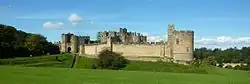 Photographie donnant une vue d'ensemble du château d'Alnwick, avec ses longs remparts de pierre, dans la campagne anglaise verdoyante, par beau temps.