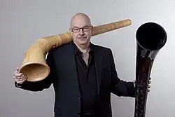 Armin Bachmann&nbsp;(de) posant avec deux parties de cor des Alpes, l'une en bois et l'autre en fibres de carbone.