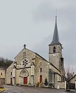Façade à collatéraux et trois portes rouges, le clocher carré à longue flèche est en retrait sur le transept.