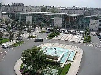 Photographie d'une gare de verre, un rond-point avec une fontaine au premier plan.