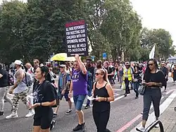 Une foule d'hommes et de femmes marchent dans une rue urbaine, avec des arbres en arrière-plan. Certaines personnes portent des pancartes