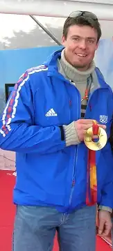 Photographie d'un homme en jean et veste bleue portant l'inscription France et les anneaux olympiques, avec une médaille d'or autour du cou qu'il tient de sa main droite.