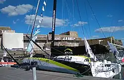 Vu de trois quarts arrière, voilier blanc et jaune amarré au ponton. En arrière-plan la base de sous-marins de Lorient.