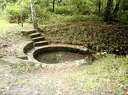 Vue d'une source entourée d'une margelle en pierre formant bassin et vers laquelle descend un petit escalier.