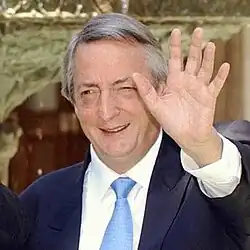 Une homme aux cheveux gris, en costume bleu sombre salut de la main gauche.