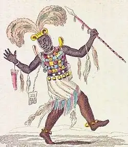 personnage africain portant une grande coiffe dorée avec des plumes d'autruche, une lance, une armure colorée avec plusieurs fétiches, un pagne et divers bijoux