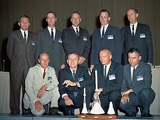 Photographie en couleur du groupe groupe d'astronautes 2 : les hommes en costume sont alignés sur deux rangées avec au premier plan des maquettes de vaisseaux spatiaux.