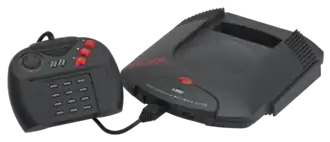 Photo de la console Jaguar à laquelle est branchée une manette de jeu, composée d'une croix directionnelle, deux boutons centraux et trois gros boutons rouges à droite, et de douze boutons dans sa partie centrale