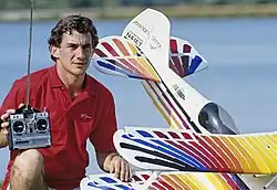 Photo d'Ayrton Senna en polor rouge qui prend la pose avec un avion miniature téléguidé, télécommande à la main.