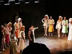 Photographie d'une scène de théâtre où deux groupes d'acteurs costumés de façon médiévales se font face. Au milieu d'eux un homme montre le groupe de droite de façon accusatrice.