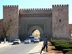 Route passant sous une porte monumentale à arc outrepassé, flanquée de deux imposantes tours symétriques, et se poursuivant sur une muraille.