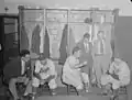 Deux joueurs des Royaux de Montréal et un joueur des Dodgers de Brooklyn assis sur des tabourets dans le vestiaire, avec trois autres hommes en costume, en 1946.
