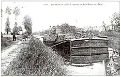 Carte postale noir et blanc du début du siècle. Un bateau, de type bâtard nivernais, une embarcation pouvant transporter 120 tonnes de marchandises, est amarré au canal. Il est chargé de rondins de bois. Des ouvriers, à gauche, posent à côté de leur brouette spécifique pour le transport du bois, une brouette n’étant pas fermée sur le côté et pouvant transporter les rondins posés transversalement. Plusieurs planches posées transversalement entre le chemin de halage et le bateau font office de passerelle.