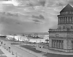 Photographie d'un grand monument de base cubique surmontée par une partie cylindrique à colonnades au sommet de laquelle se trouve un toit conique. Plusieurs cuirassés sont visibles dans le fleuve à l'arrière-plan.