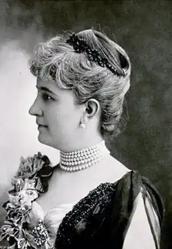 Vue rapprochée, de profil, d'une femme aux cheveux courts et blonds, portant un diadème et un collier de perles.