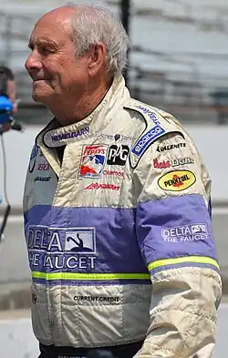 Photographie d'un homme blanc, âgé, de profil, en combinaison de pilote grise et violette, vue de profil, en plan américain.