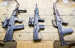 photographie en couleur montrant trois fusils d’assaut accrochés côte à côte à la verticale sur un mur en bois