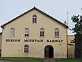 La façade d'un bâtiment aux murs ocre avec six fenêtres en ogive. Les mots « BRECON MOUNTAIN RAILWAY » y sont écrits en grandes lettres marron.