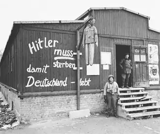 Photo noir et blanc de l'entrée d'une baraque en bois dans le camp de concentration de Buchenwald. Au centre de la photo, près du mur de la baraque, se dresse une potence à laquelle pend un pantin à l'effigie d'Adolf Hitler. Sur le mur de la baraque est peinte l'inscription en allemand : « Hitler muss sterben damit Deutschland lebt ». À droite, un homme, en tenue sombre, se tient debout dans l'encadrement de la porte, au sommet de l'escalier en bois qui mène à l'entrée. Un soldat américain pose debout à côté de l'escalier.