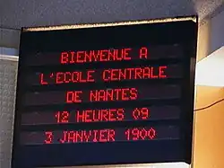 Tableau électronique dans une école qui affiche « Bienvenue à l'école centrale de Nantes, 12 heures 09, 3 janvier 1900 ».