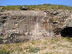 Mur d'un blockhaus éclaté en plusieurs endroits (impacts d'obus visibles). Herbe sur le dessus.