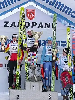Quatre jeunes femmes en tenue de sport et dossard numéroté de compétition tenant des skis de saut verticalement d'une main, dont trois sur un podium chacune sur une marche, tenant chacune une coupe en métal doré d'environ 50 centimètres, et une à leur gauche, sur fond pavoisé de marques de sponsors et marqué « ZAKOPANE »