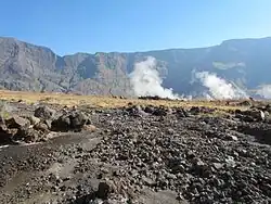 Photographie couleur d'une plaine caillouteuse entourée de falaises rocheuses. Des fumées blanches s'échappent du sol.